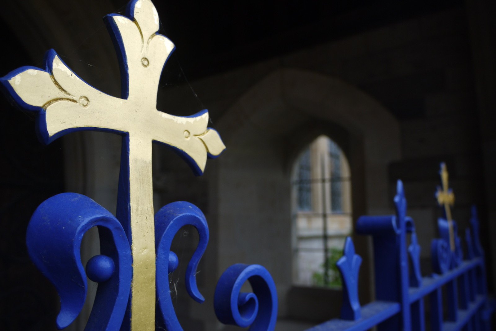 Close-up of a blue and gold cross on a church gate, with a stained glass window in the background.