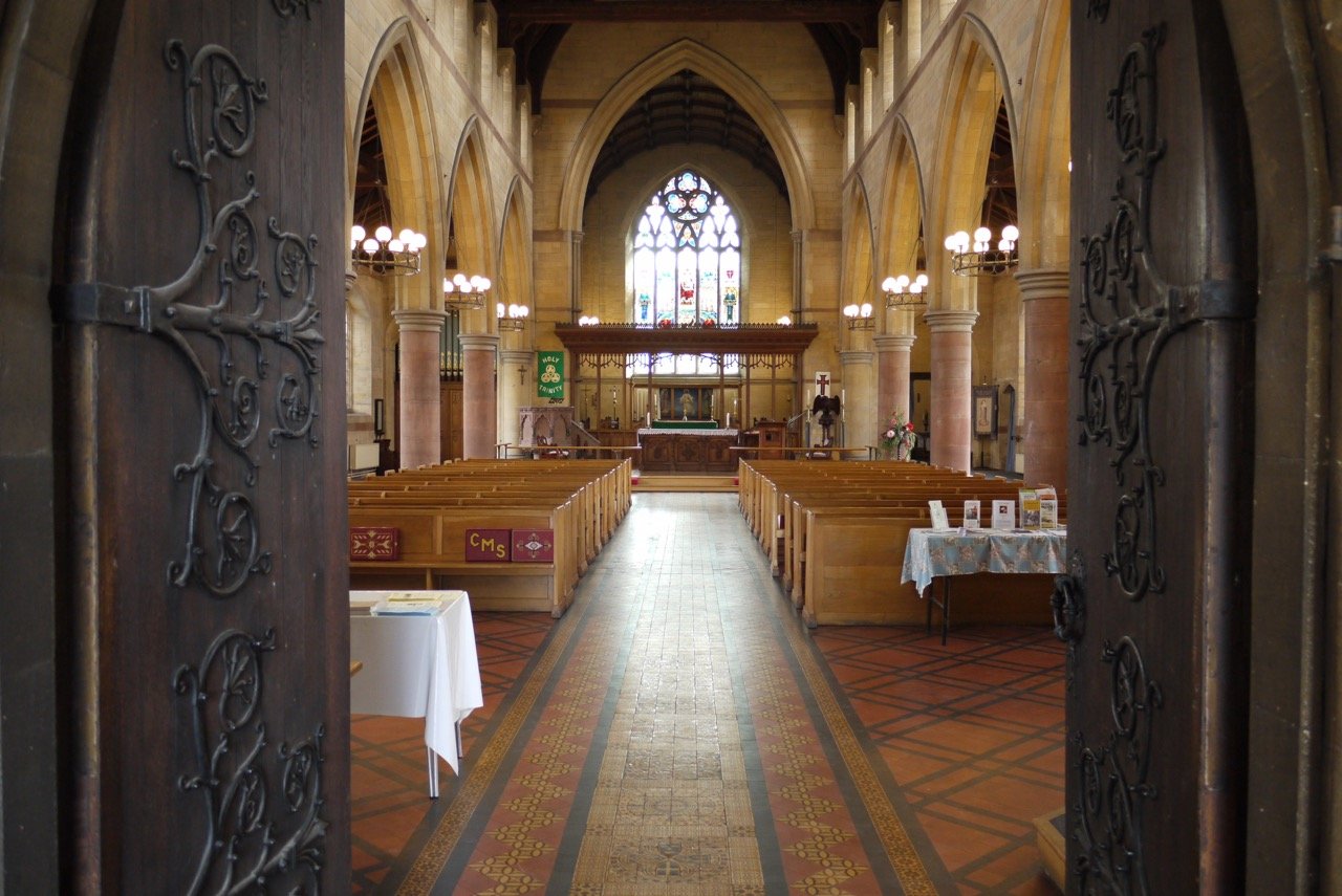 View through carved wooden doors into the church nave, with pews, covered tables, and the altar beyond.
