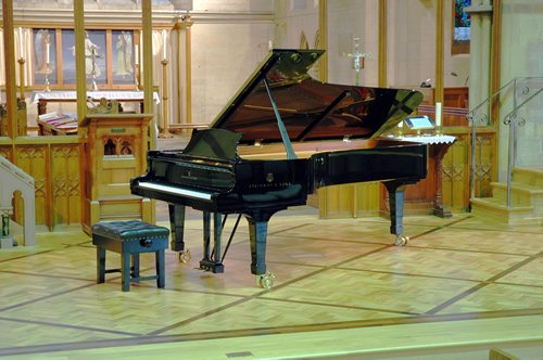 A grand piano set up in a church for a recital, with wood panelling and stained glass behind.