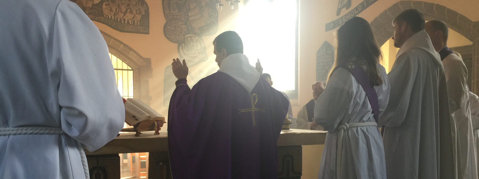 Clergy in white and purple vestments face the altar during a service, seen from behind.