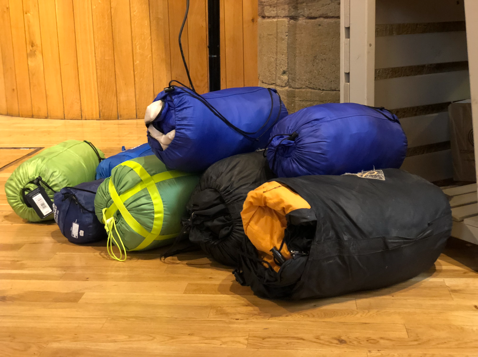 A pile of stuffed sleeping bags and duffel bags on a wooden floor.