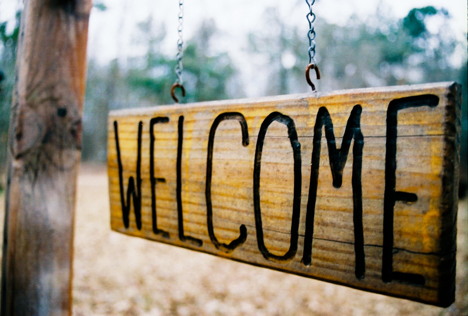 A wooden sign reading 'Welcome' hangs by chains from a post, with trees in the background.