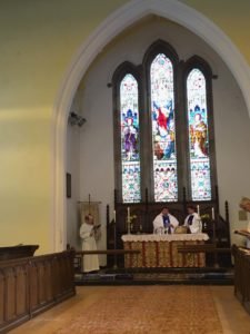 Clergy stand at the altar of a church beneath a tall three-light stained glass window.