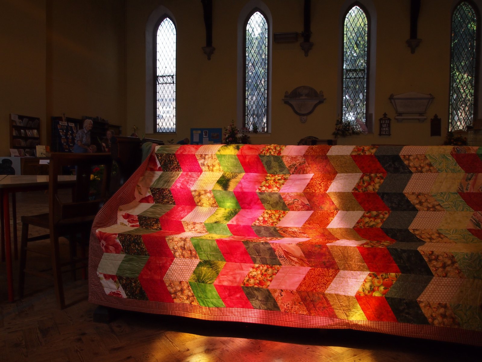 A patchwork quilt draped over a stand in a church, lit by coloured light from a stained glass window.