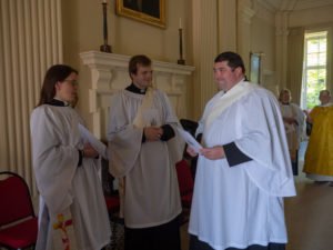 Three clergy in white robes talk together in a church during an ordination service.