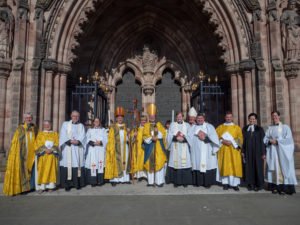 A group of clergy in vestments pose together outside the west door of a cathedral.