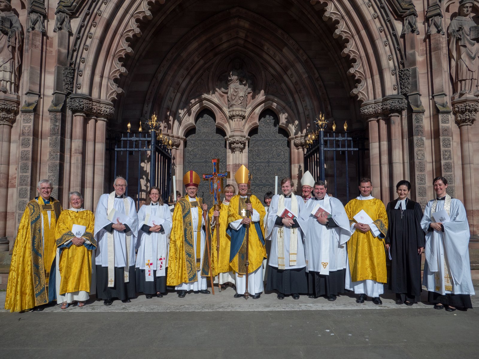 A group of clergy in vestments pose together outside the west door of a cathedral.