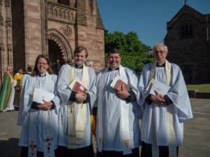 Four clergy in white robes stand together outside Hereford Cathedral on a sunny day.