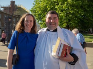 A woman in a blue dress and a priest in white robes smile together outside Hereford Cathedral.