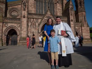 A woman, a young boy, and a priest in white robes stand together outside Hereford Cathedral.