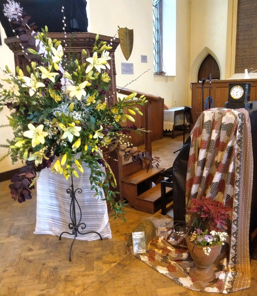 A white lily arrangement displayed in a church, beside a patchwork quilt.