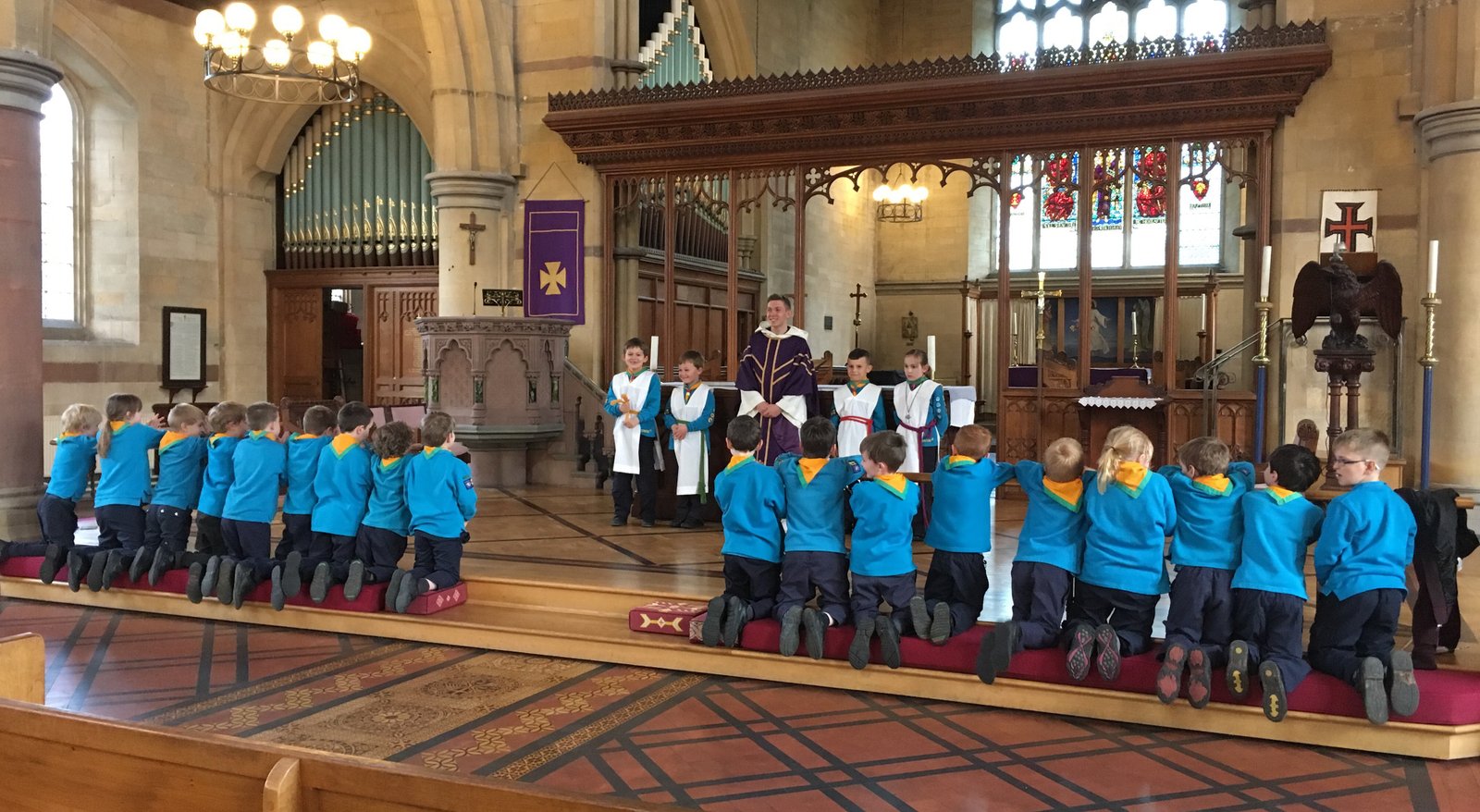 Children in blue uniforms kneeling in church pews, with clergy in purple vestments at the altar.