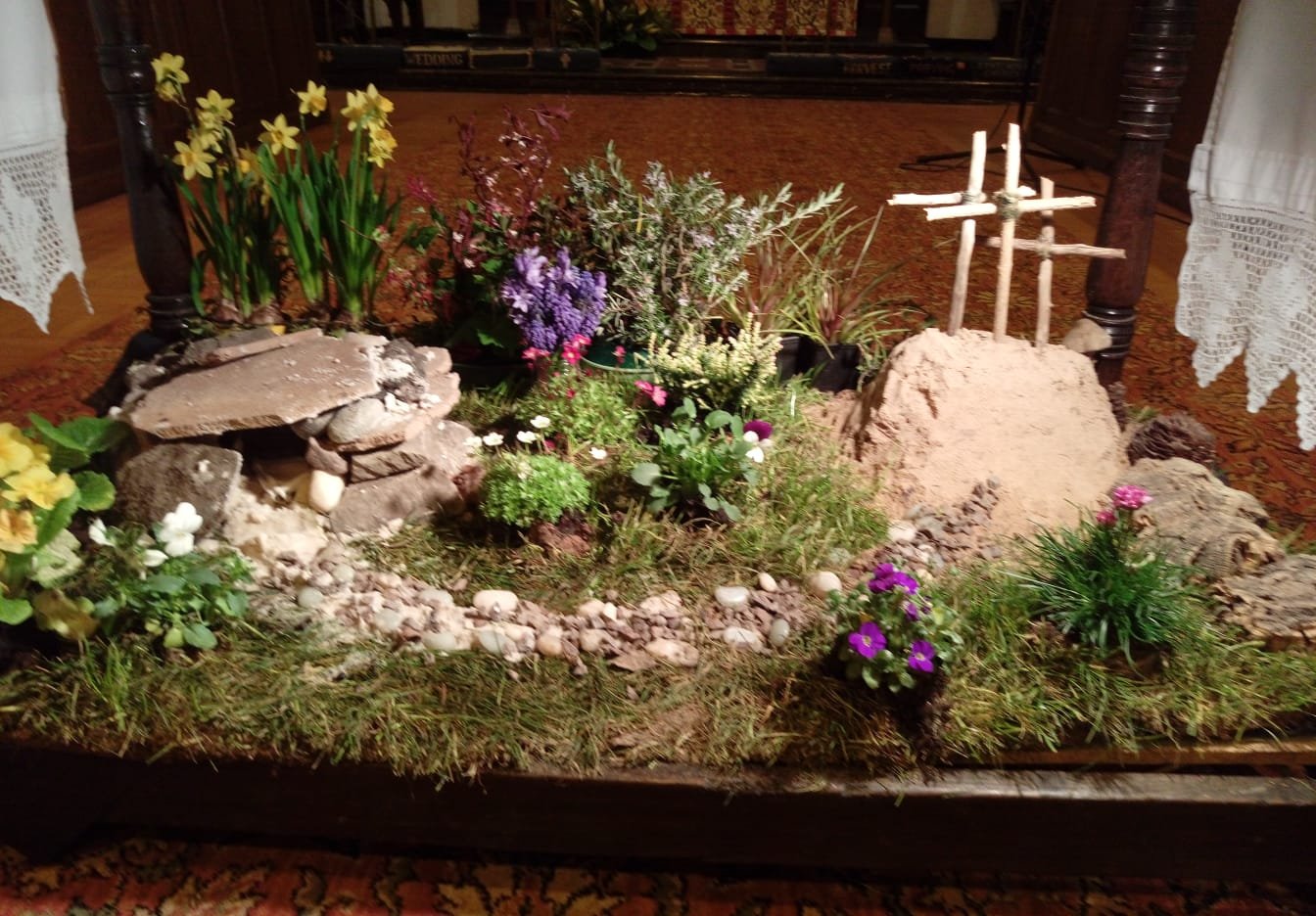 A miniature Easter garden display with moss, flowers, small stones and three wooden crosses on a hill.