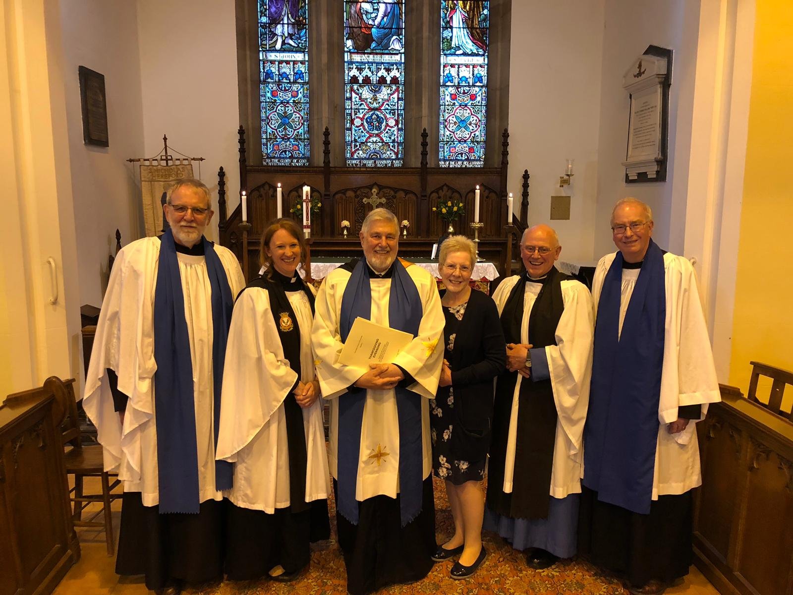 Six clergy in white and blue robes stand for a group photo in front of the altar and stained glass window.