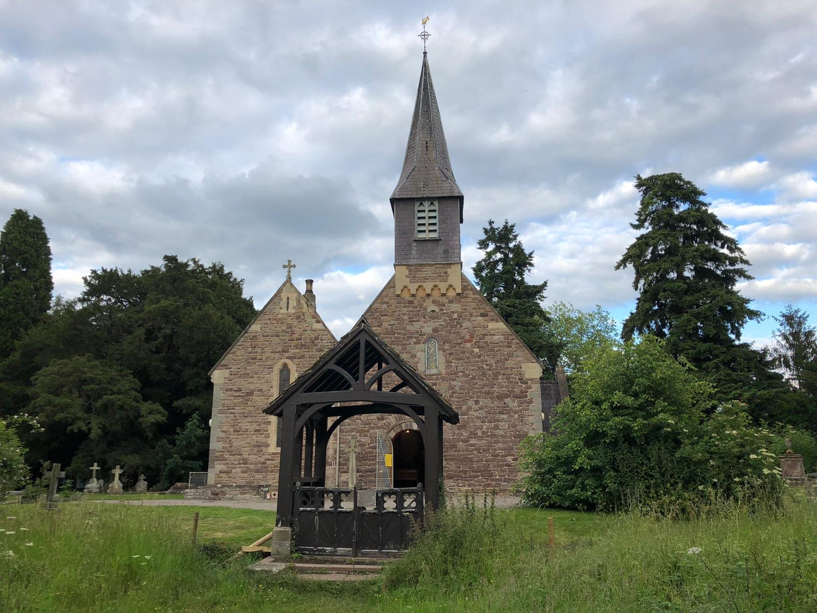 A small stone church with a bell tower and spire, seen beyond a wooden lychgate surrounded by trees and long grass.