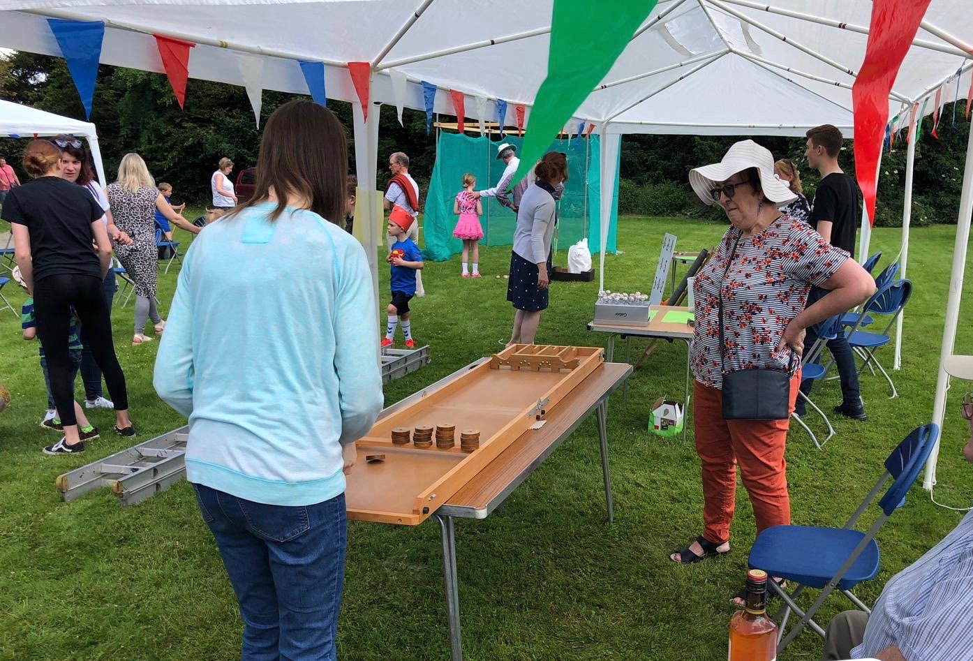 Fete-goers play a table skittles game under a marquee, with bunting and gazebos in the background.