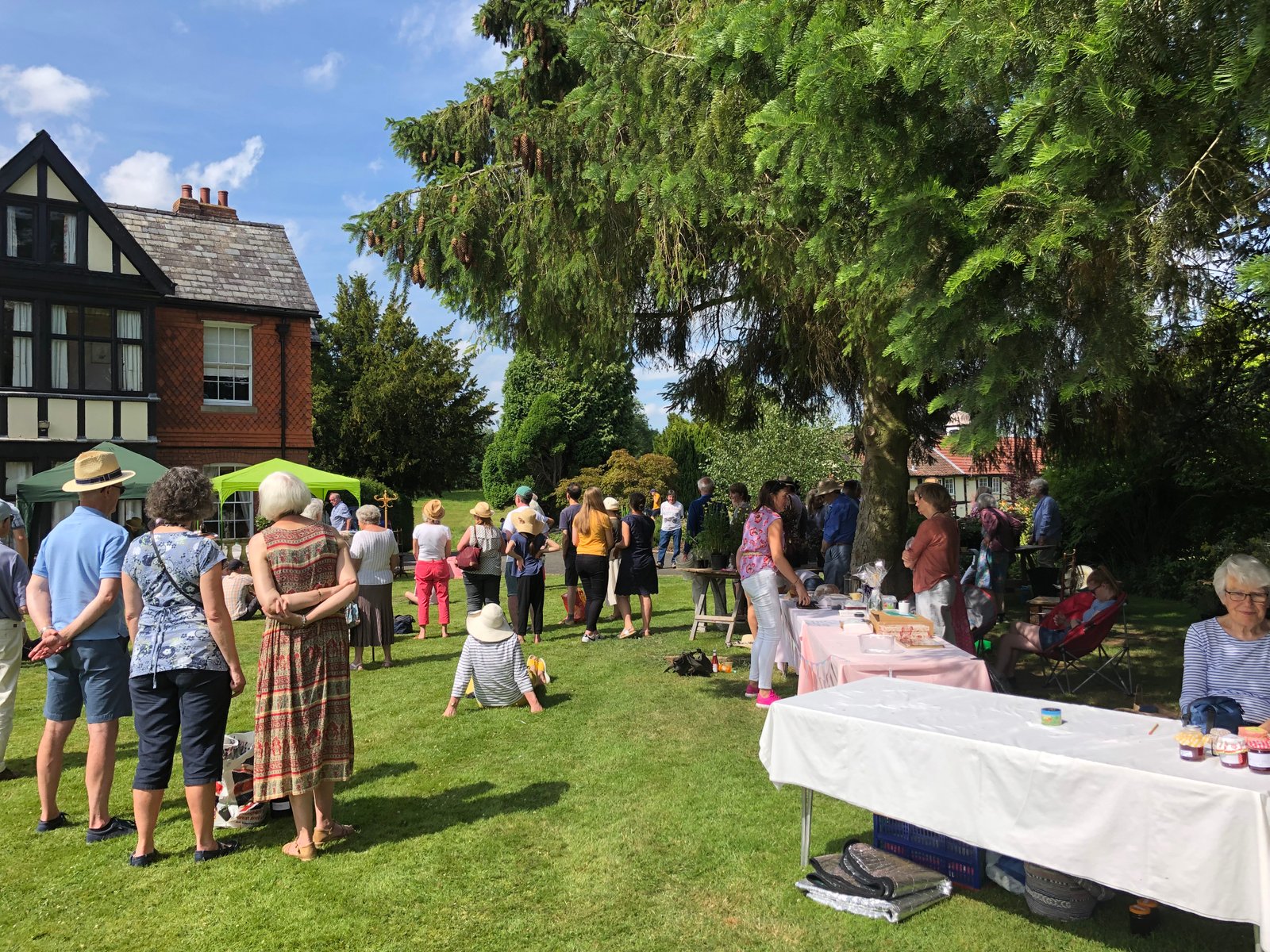 Parishioners gather on the lawn at a summer garden party, with stalls set up near a Tudor-style house.