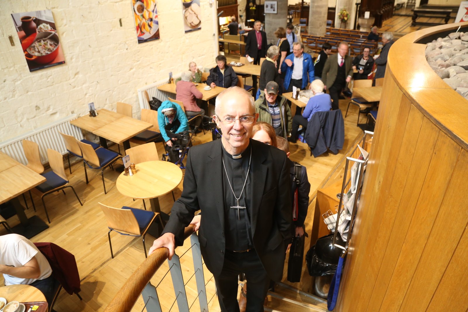The Archbishop of Canterbury walks down a staircase into a busy cafe area with people seated at tables.