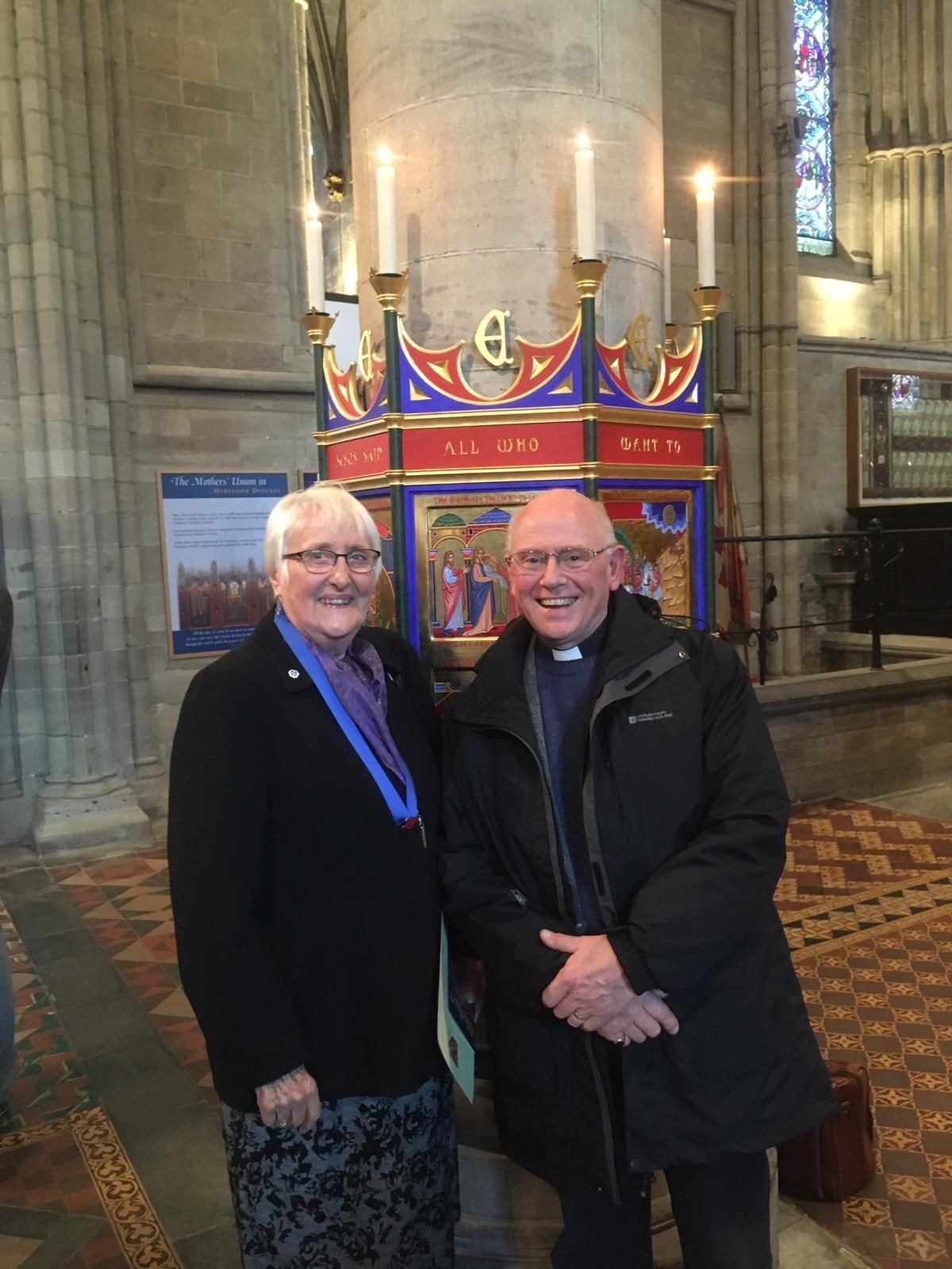 A clergyman and a woman wearing a ceremonial medallion stand together in front of a lit altar in a cathedral.