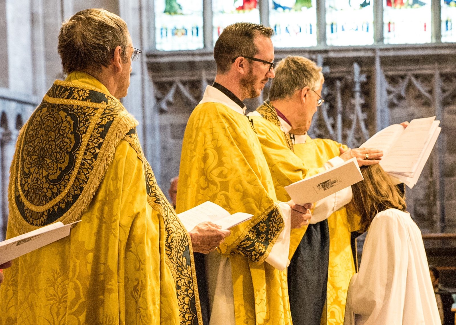Clergy in gold vestments stand together during a service, with stained glass windows behind them.