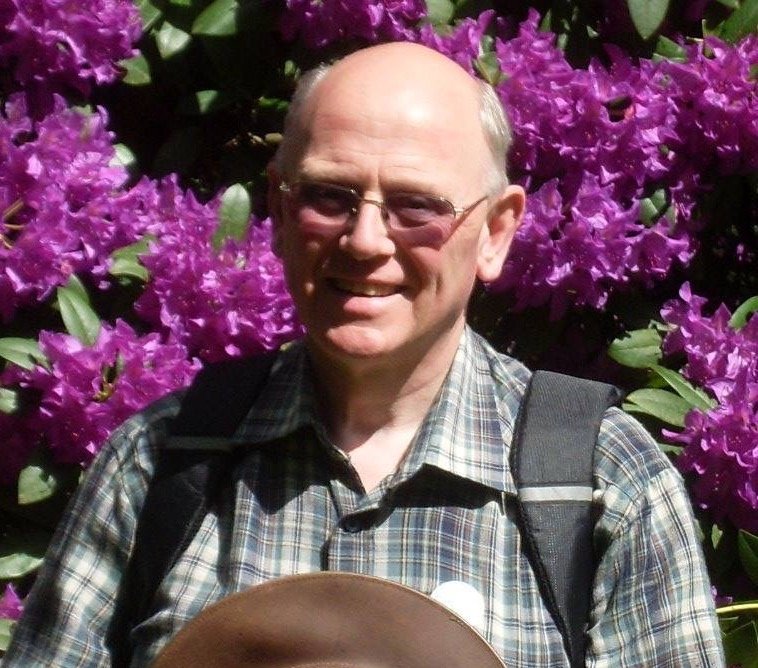 Portrait photo of a man with glasses standing in front of purple rhododendron flowers.