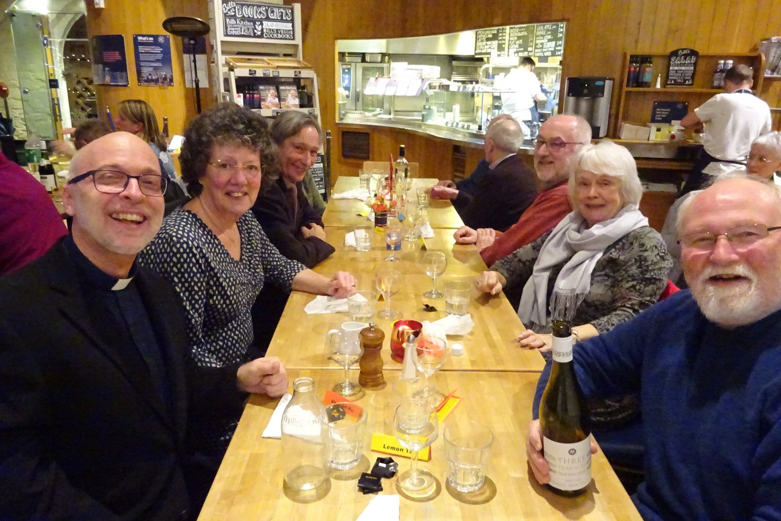 A group of friends smile for the camera around a table set with glasses at a pub meal.