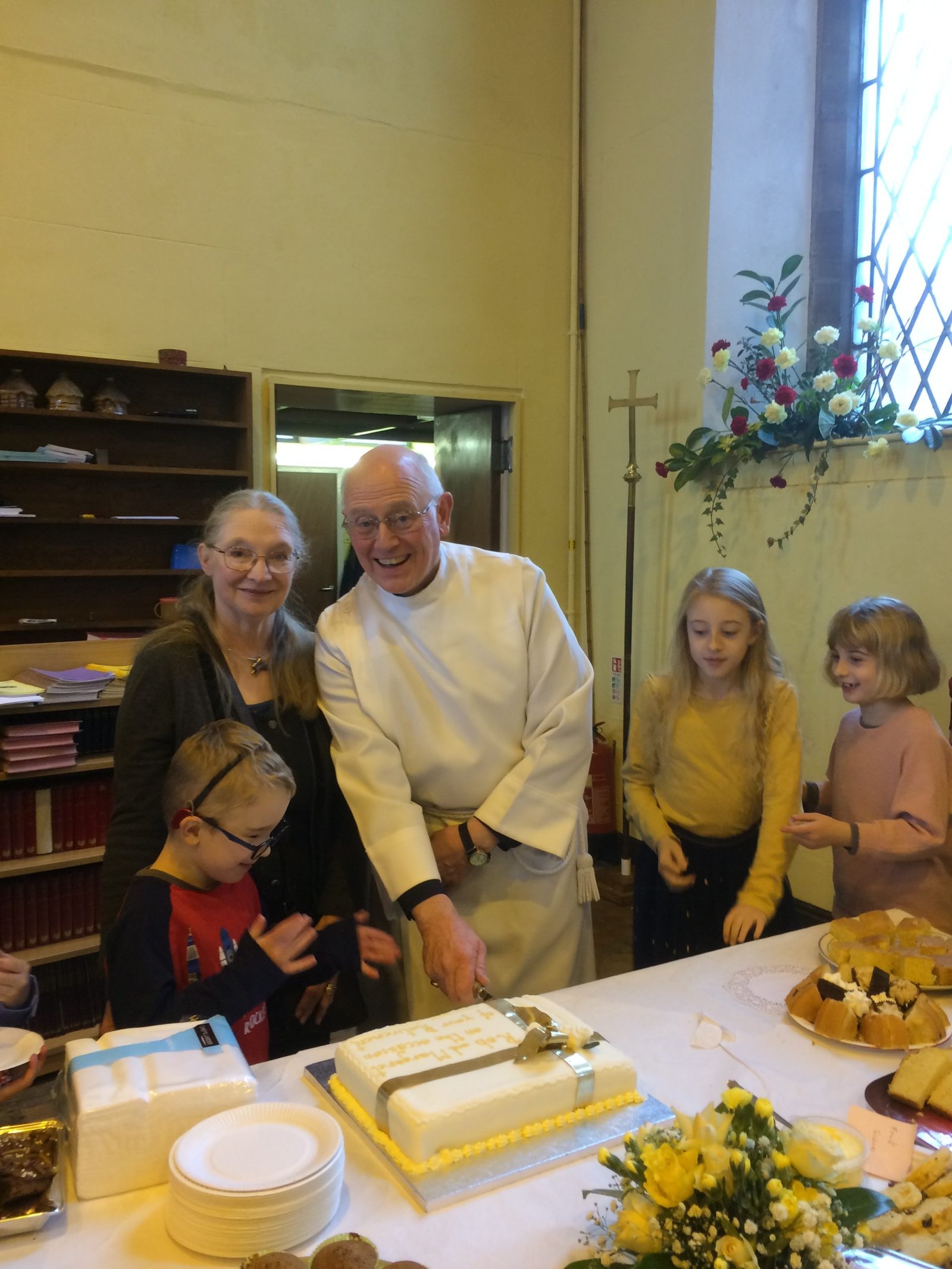 A vicar cuts a cake at a table with family and children looking on in a church hall.