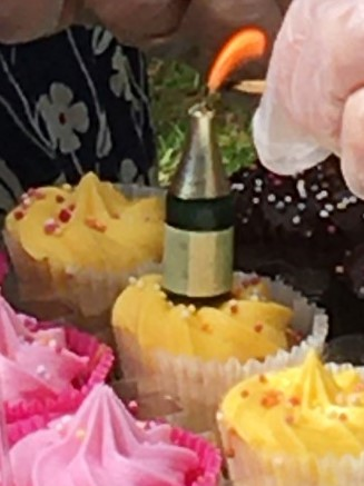 A lit sparkler shaped like a bottle stands among iced cupcakes at a birthday celebration.