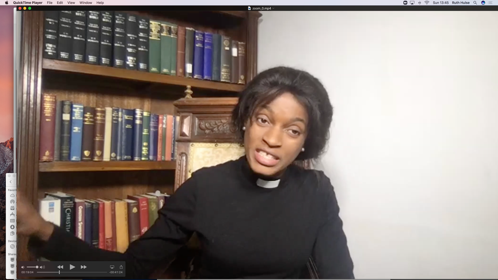 Screenshot of a woman in a clerical collar speaking during an online service, seated in front of a bookshelf.