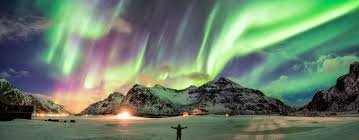 The aurora borealis glows green and purple over snow-capped mountains, with a person standing with arms raised below.