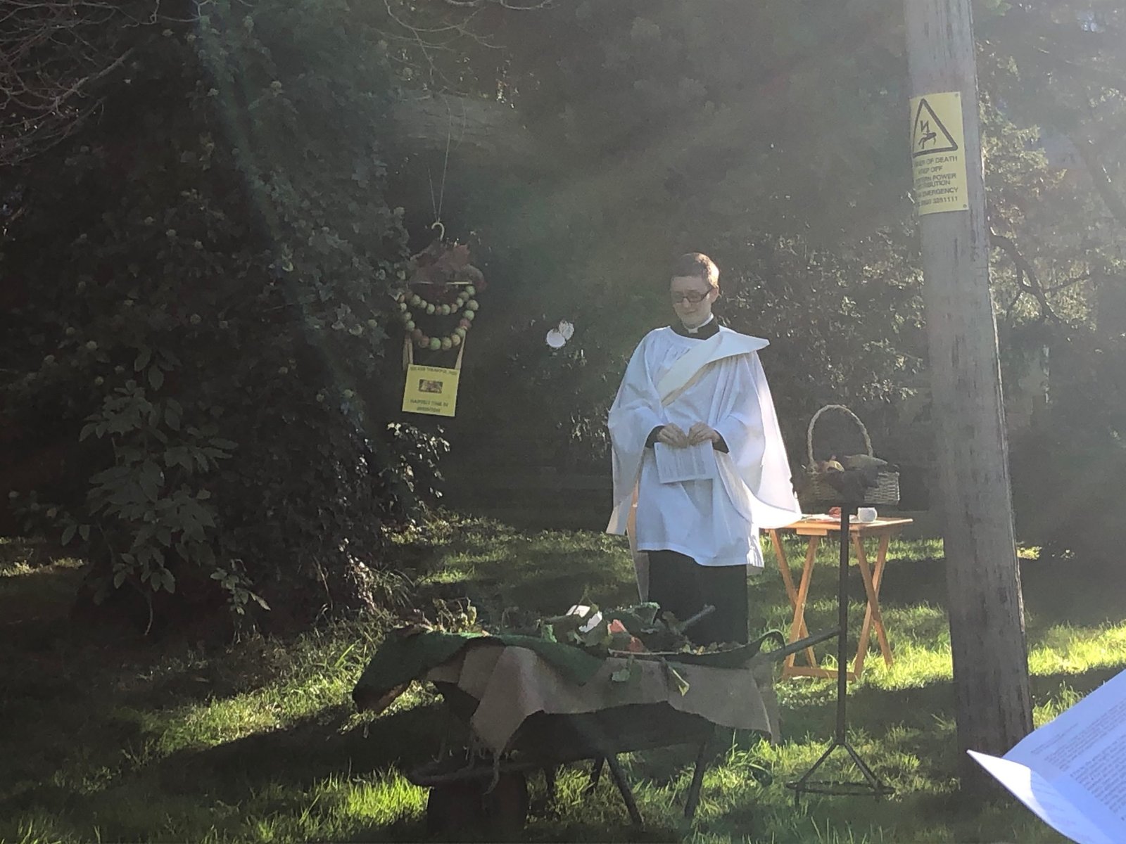 A priest in white robes stands outdoors in dappled sunlight beside a small table set with flowers.