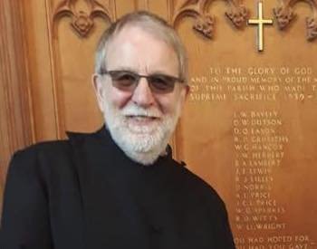 Portrait photo of a priest with glasses and a white beard, standing in front of a war memorial plaque inside a church.