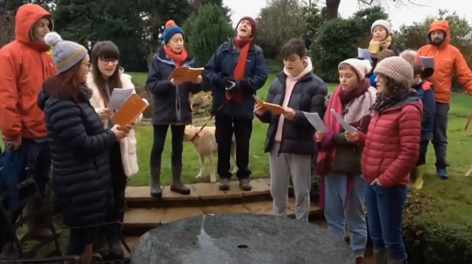 Carol singers gather outdoors in winter coats and hats, holding song sheets, with a dog beside them.