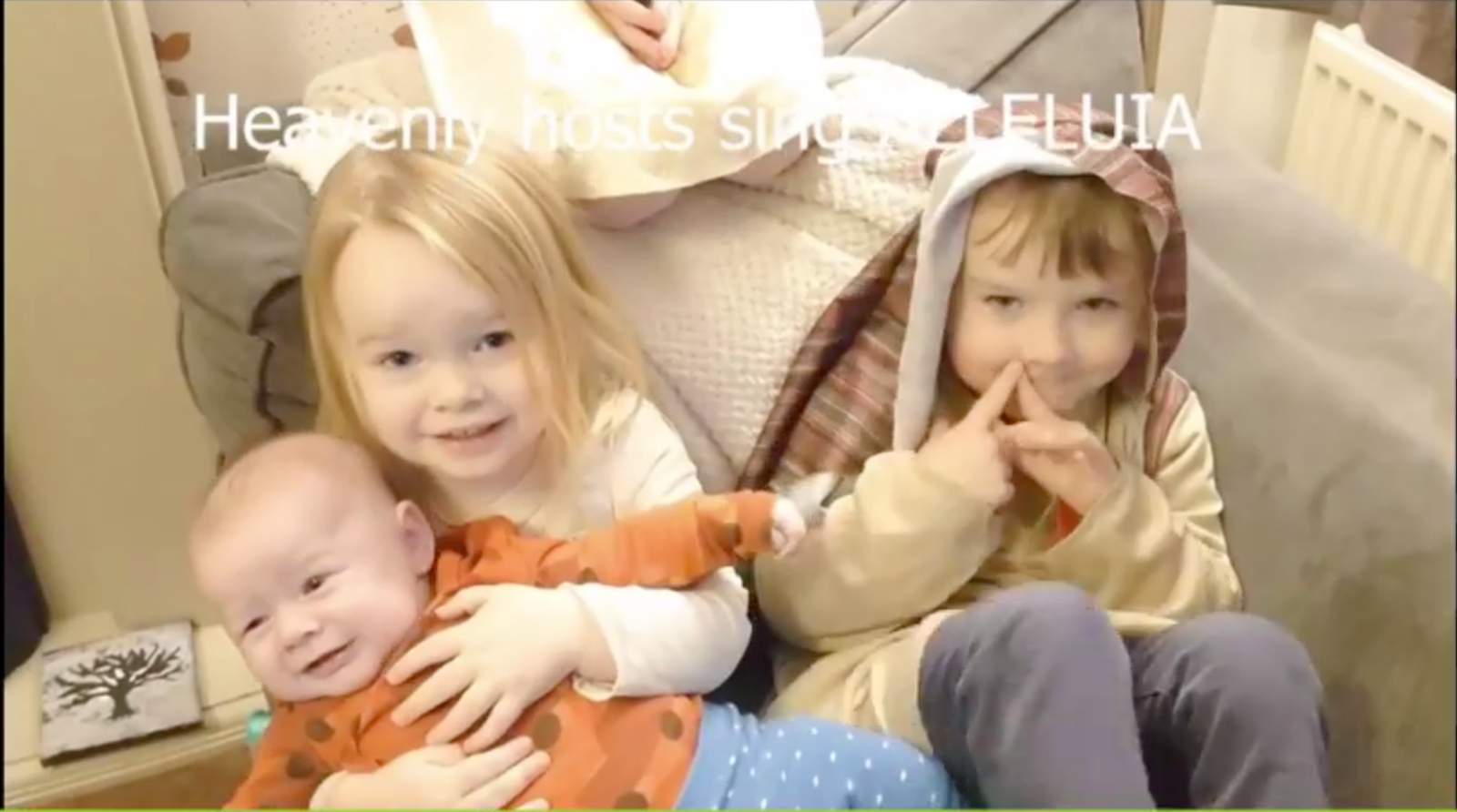 Video call screenshot of three young children on a sofa, with text overlay 'Heavenly hosts sing Alleluia'.