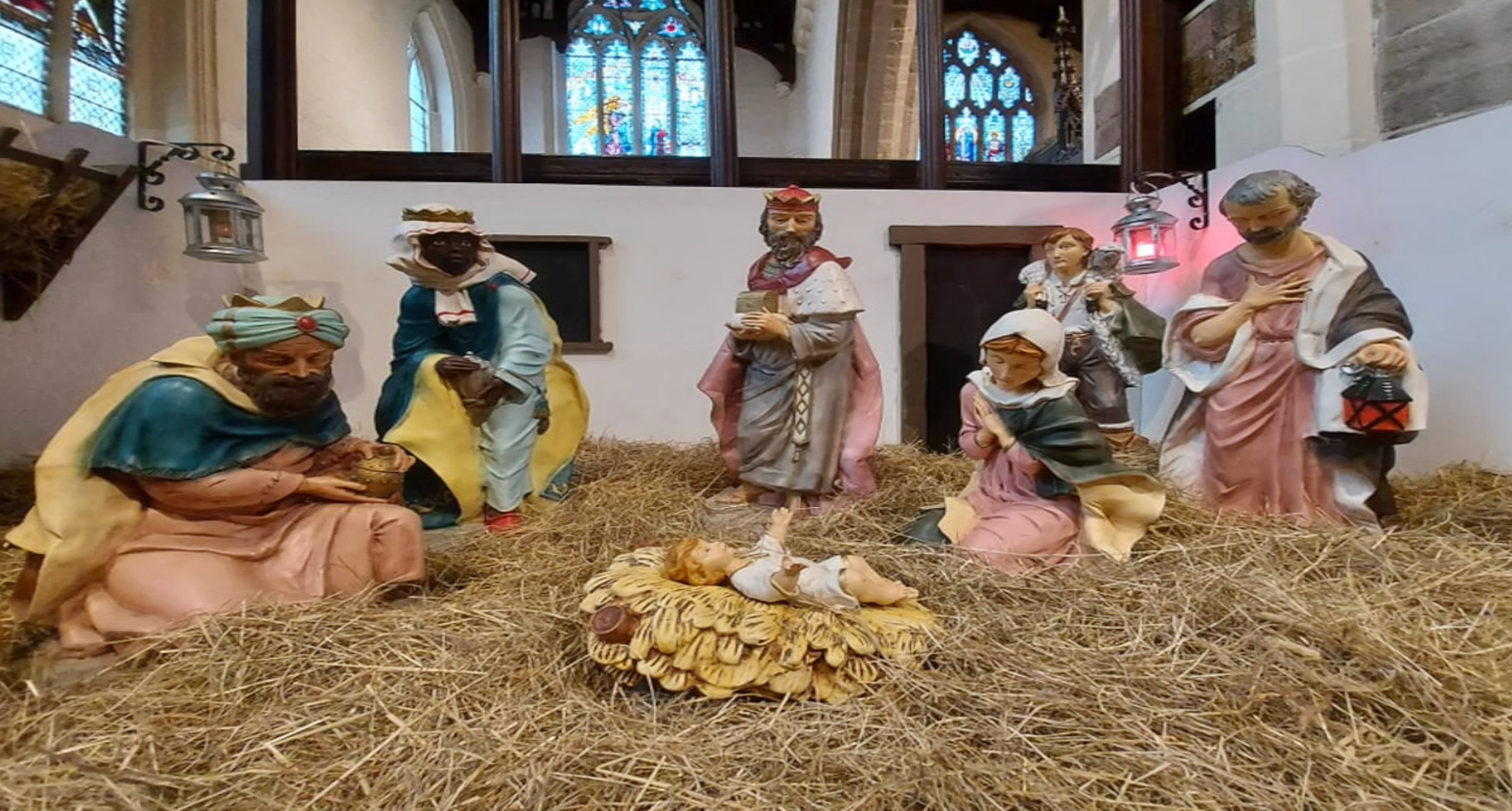 Nativity figures depicting the Three Kings and shepherds gathered around the manger, set inside a church with stained glass windows behind.