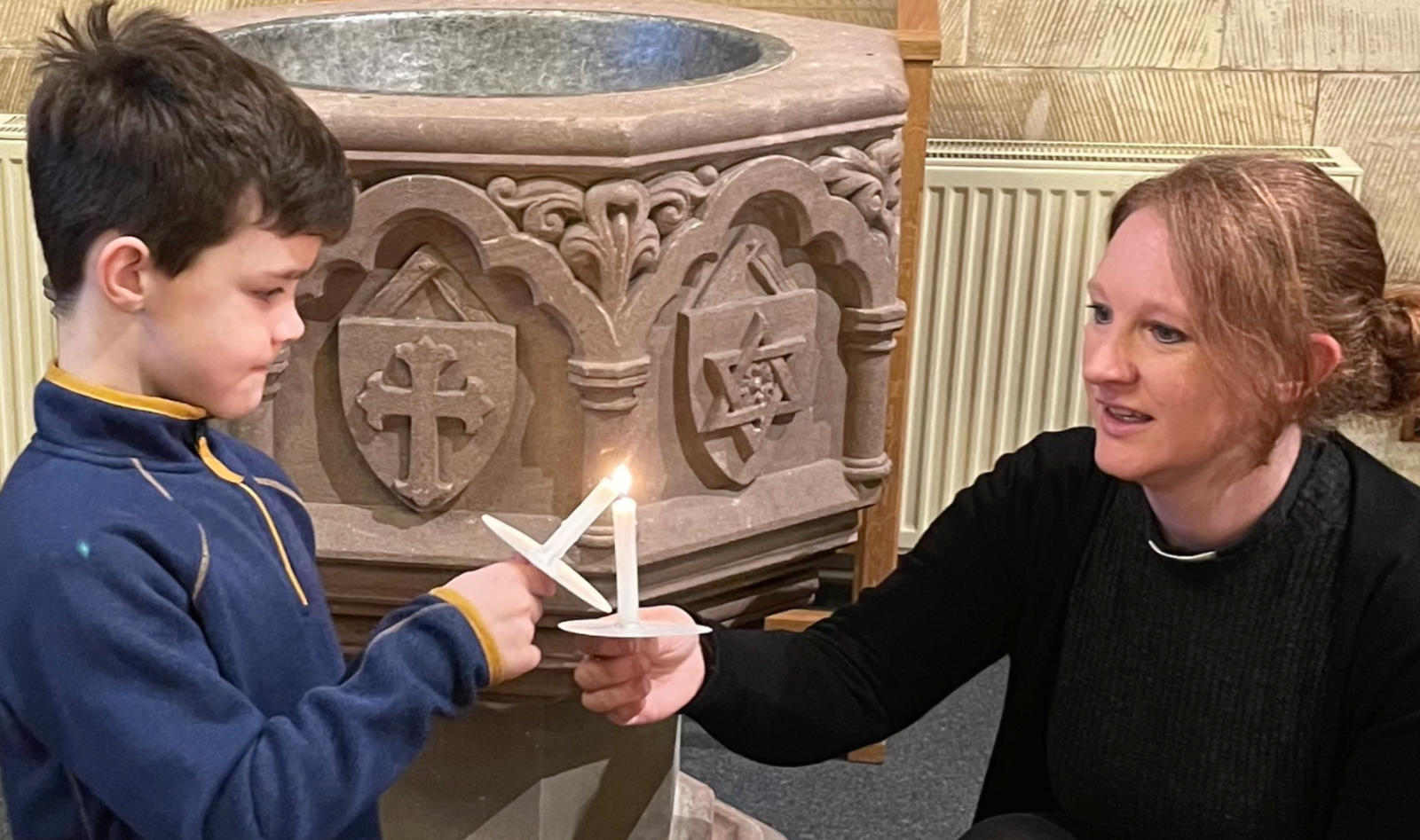 A woman helps a boy light a candle beside a carved stone font in church.
