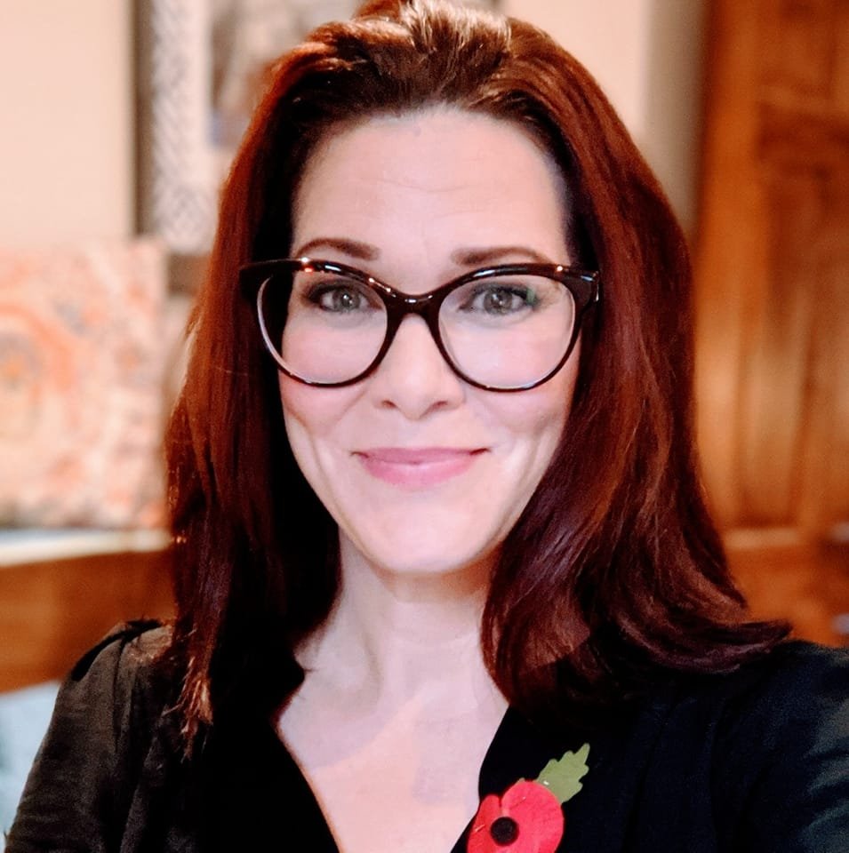 Portrait photo of a woman with red hair and glasses, wearing a poppy pin.