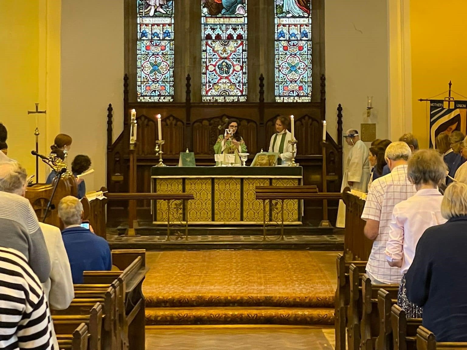 Clergy stand at the altar beneath a stained glass window as the congregation looks on during a Eucharist service.