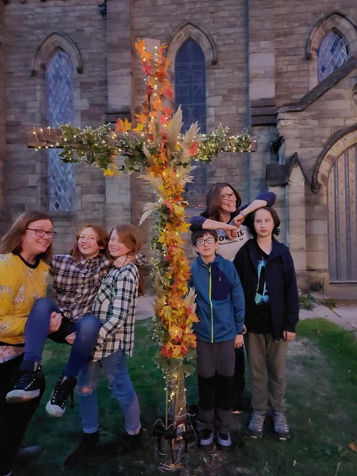 Children and an adult stand around a large cross decorated with autumn leaves outside a church.
