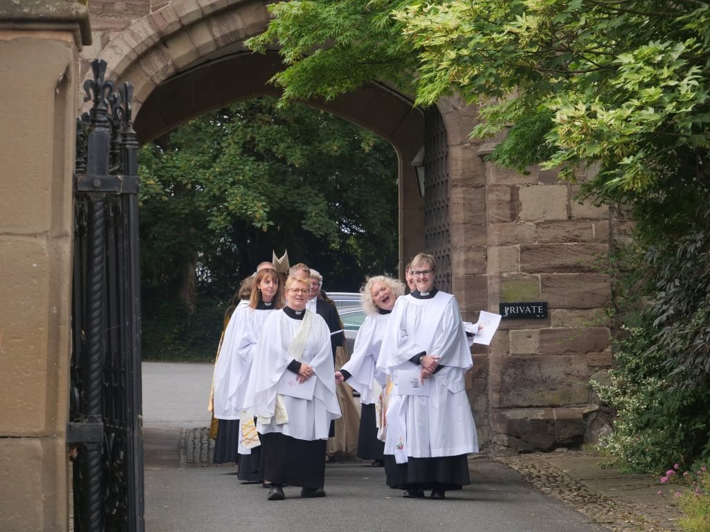 Clergy in white robes walk through an arched stone gateway at Hereford Cathedral.