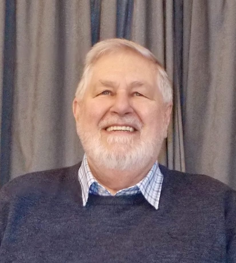 Malcolm Robertson, a smiling man with a white beard, wearing a blue sweater and checkered shirt.