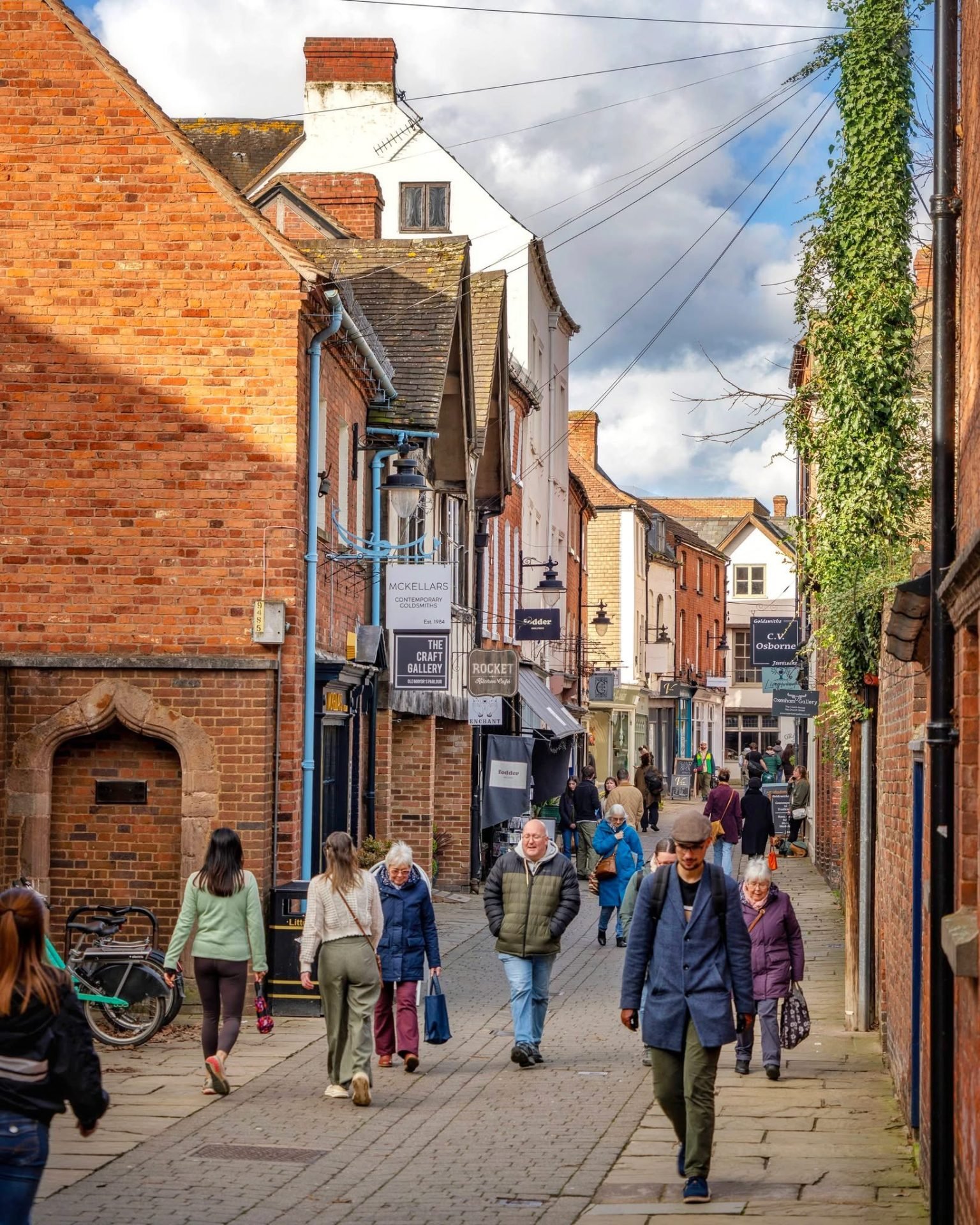 People stroll down a charming brick-lined shopping street with ivy-covered walls.