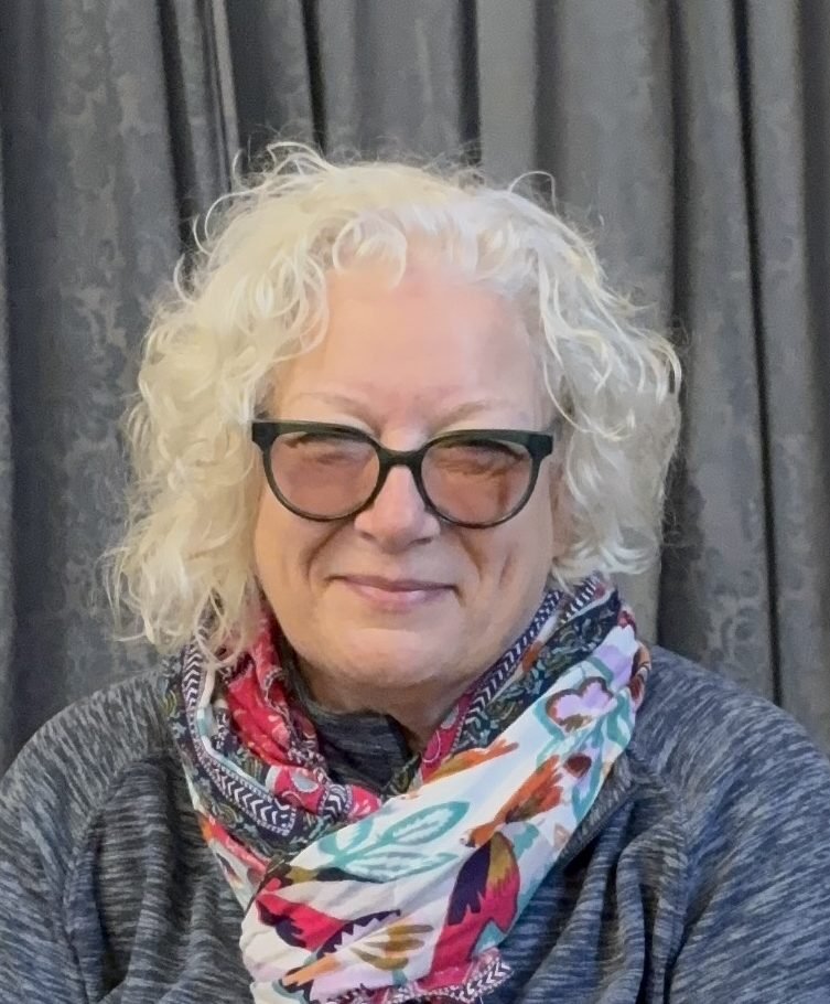 Melody Carter, a woman with curly blonde hair and glasses, smiles wearing a colorful scarf.