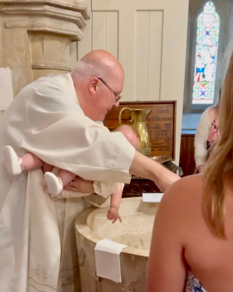 A priest in white vestments holds an infant dressed in a white gown over a stone baptismal font during a christening inside a church.