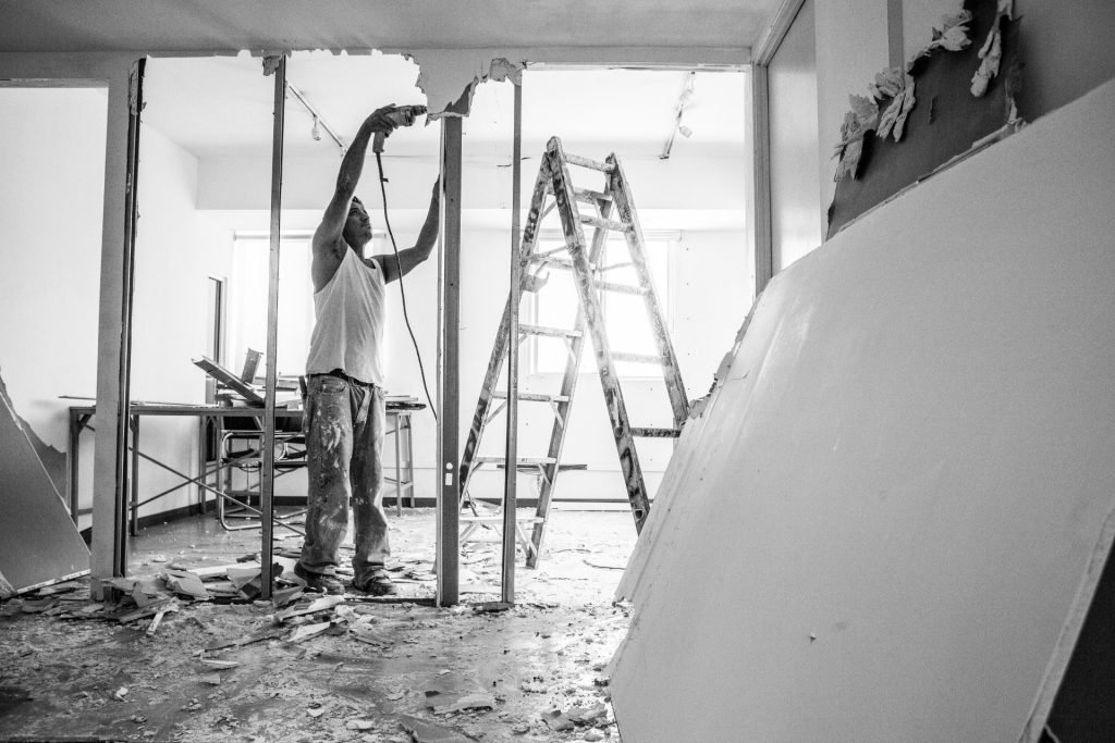Man demolishing wall with drill during renovation, ladder nearby
