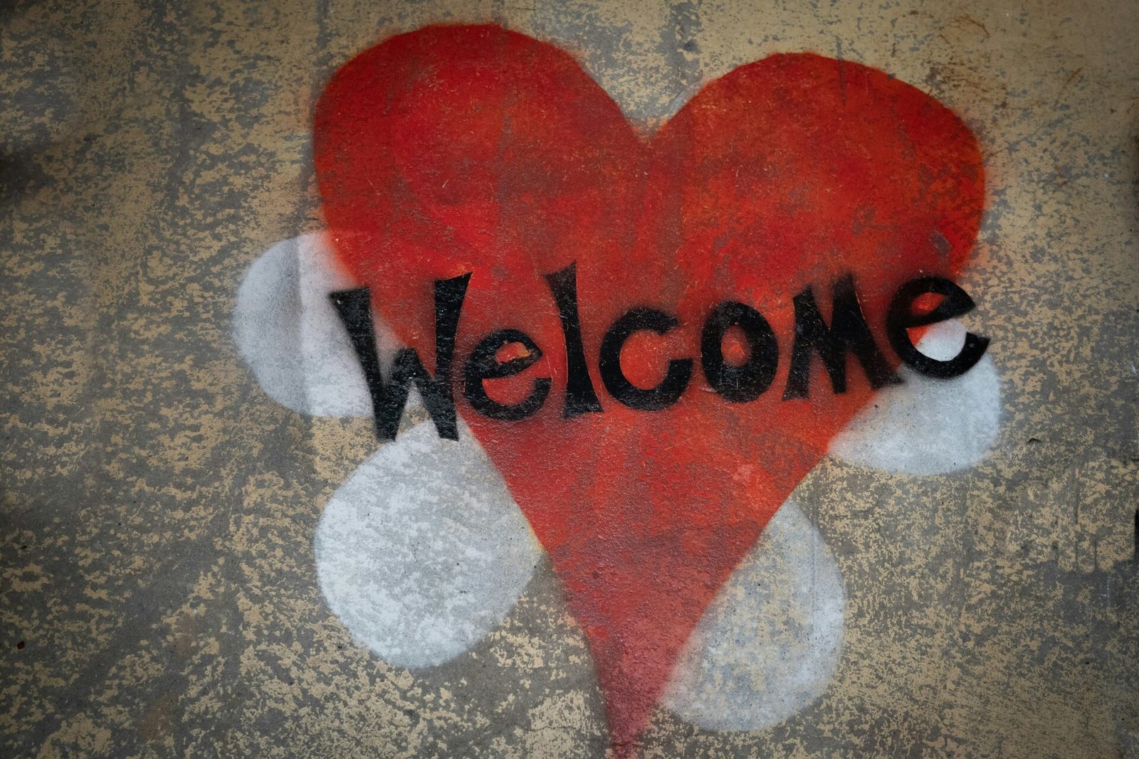 A red spray-painted heart on a textured concrete ground with the word "Welcome" stenciled across the front in black letters.