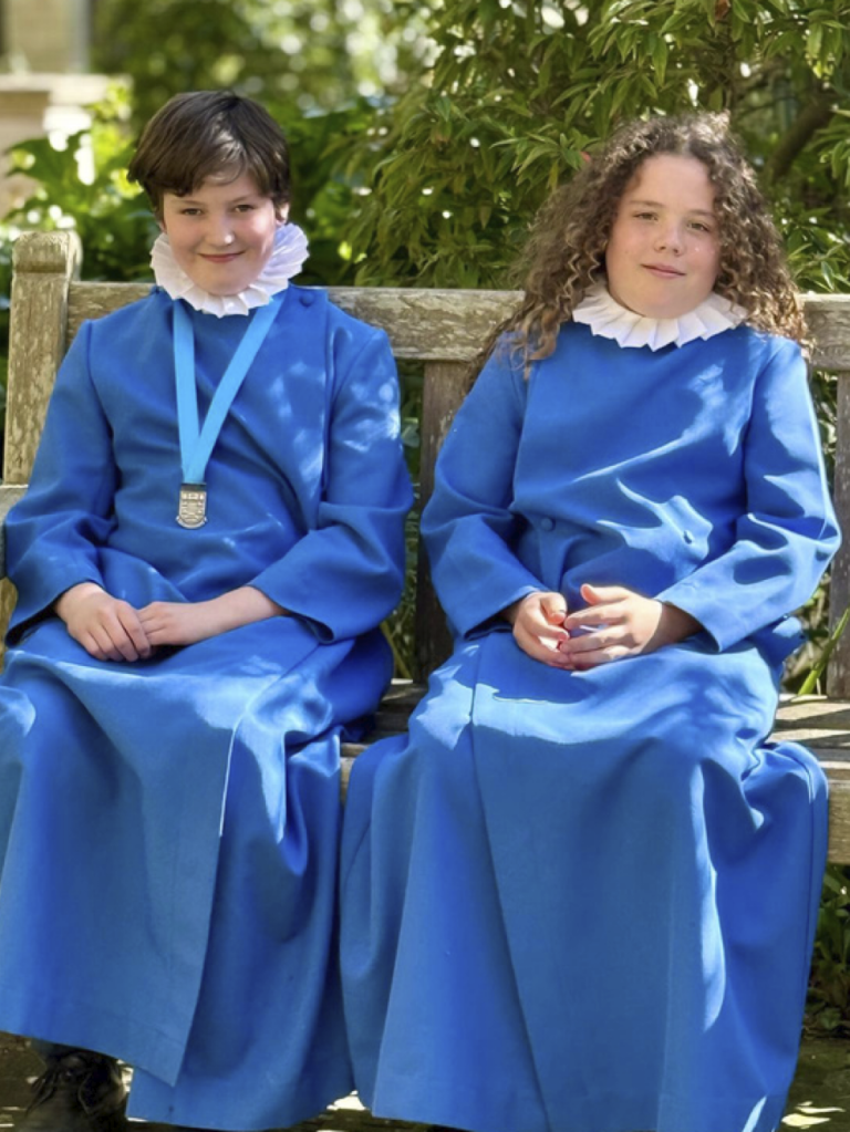 Two choir boys in blue robes with white ruffs sit on a bench.