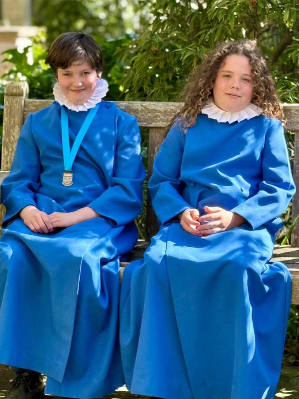 Two choir boys in blue robes with white ruffs sit on a bench.